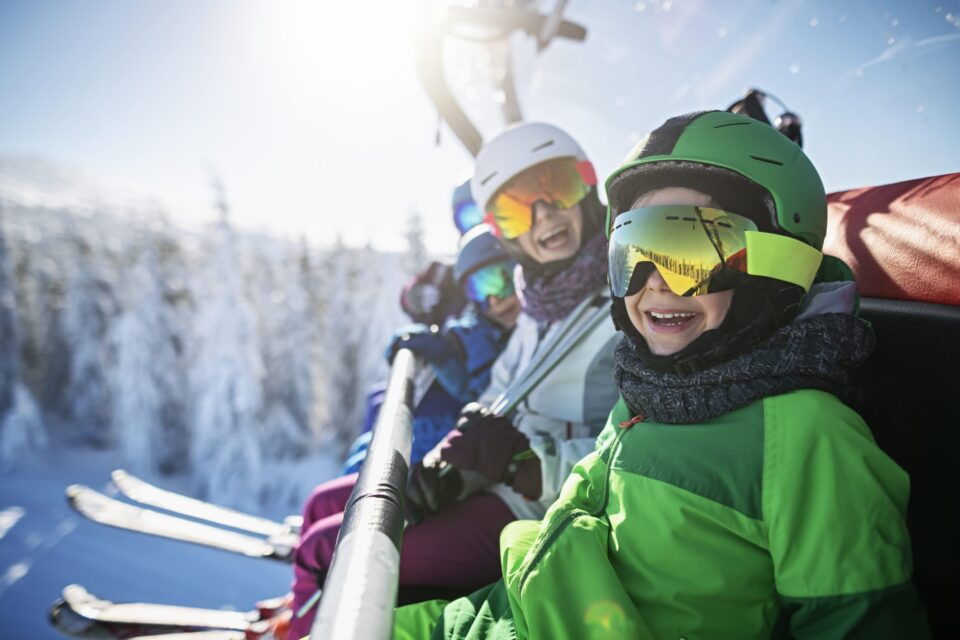 Mother skiing with kids on a sunny winter day. Family is sitting on chairlift cheering at the camera.
Nikon D850
